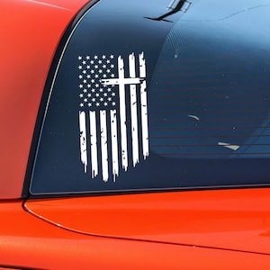 May include: A white distressed American flag decal with a white cross in the center. The decal is applied to the back window of a red car.
