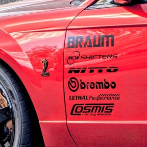 May include: A bright red car fender featuring a black cobra emblem and a series of black decals including the text BRAUM, MCW SHIFTERS, NITTO, brembo, LETHAL Performance, and COSMIS racing wheels, positioned above a black tire and gold brake caliper.