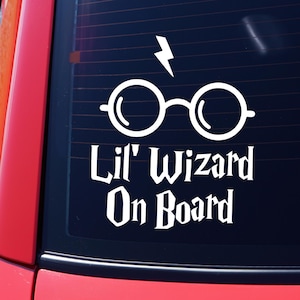 May include: White car decal featuring a pair of round glasses and a lightning bolt, with the text "Lil' Wizard On Board" in a stylized font. The decal is applied to a car window, with a red car body visible on the left.