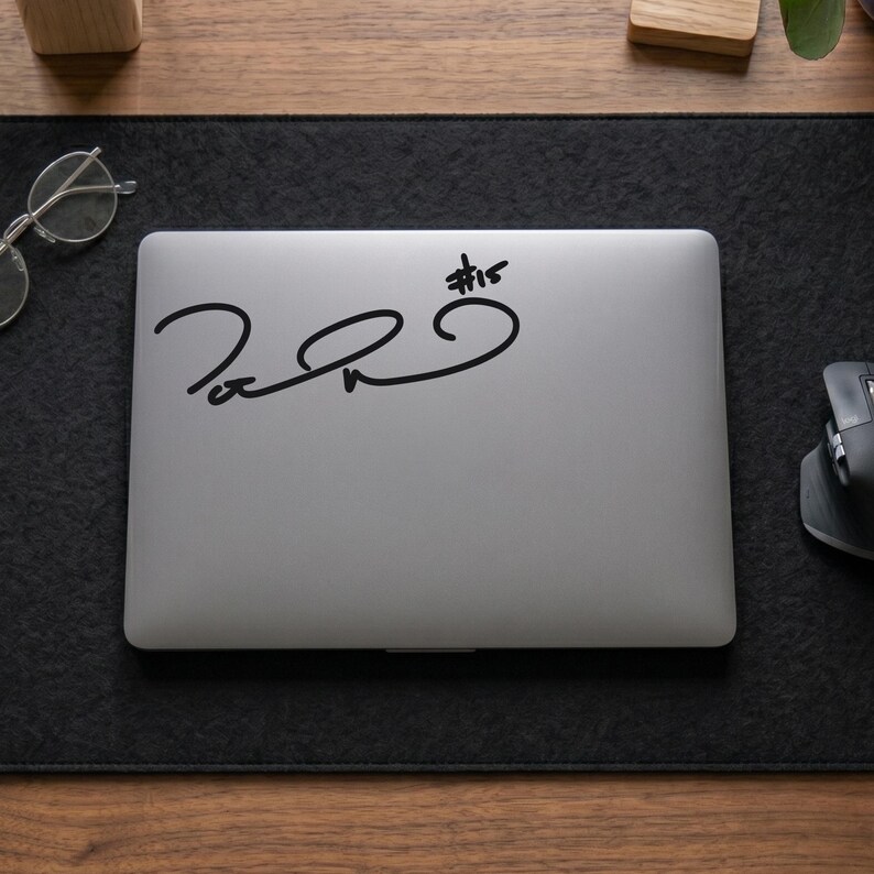 May include: A silver laptop with a black signature and the text #15 printed on the lid. The laptop is positioned on a dark gray felt desk mat, with a pair of clear-framed glasses to the left and a black computer mouse to the right.
