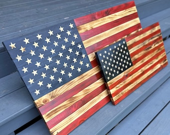 Puede incluir: Dos banderas americanas de madera, una más grande que la otra, se muestran sobre una superficie de madera gris. Las banderas están hechas de madera teñida con pintura roja, blanca y azul. La bandera más pequeña está colocada encima de la más grande.