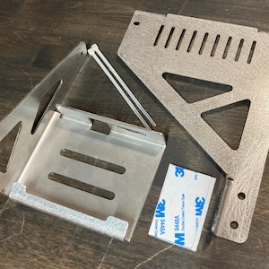 May include: Two metal brackets with cutouts and holes. The brackets are silver and have a rough texture. There are two white plastic ties and a blue 3M adhesive sticker.