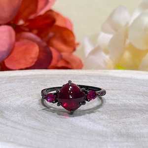 May include: A black ring with a large, square-cut, deep red gemstone at the center, flanked by two smaller, teardrop-shaped red stones. The ring has a dark, matte finish, and the gemstones are faceted.