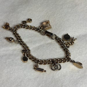 Puede incluir: Una pulsera de dijes dorada con varios dijes, incluyendo una máquina de coser, un rodillo y anillos de boda. La pulsera tiene un diseño de cadena de bordillo y un cierre. Los dijes son pequeños y detallados.