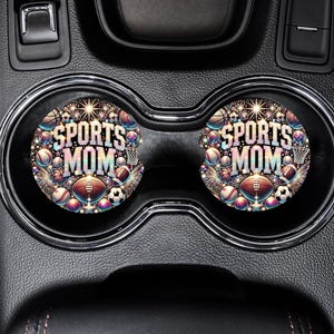 May include: Two black car cup holders with colorful round coasters that say "Sports Mom" surrounded by sports equipment like baseballs, basketballs, footballs, and soccer balls.