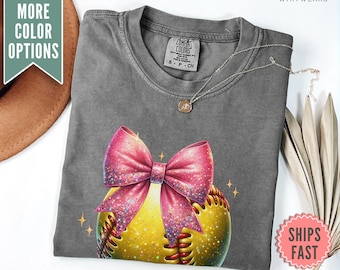 May include: A gray t-shirt with a yellow baseball with a pink bow on top. The baseball has white stitching and glitter accents. The shirt has a tag that says "Comfort Colors".