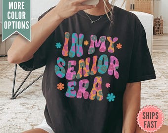 Puede incluir: Camiseta negra con un gráfico colorido que dice "In My Senior Era" con flores en las letras.