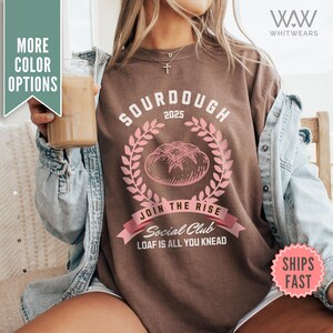 May include: Brown sweatshirt with a pink graphic design of a loaf of sourdough bread with the text "Sourdough 2025" above the bread and "Join the Rise Social Club Loaf is all you knead" below the bread.