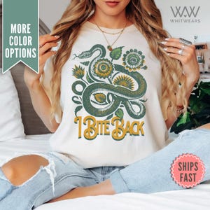 Puede incluir: Camiseta blanca con un gráfico de serpiente verde y flores amarillas. El texto "I Bite Back" está impreso en amarillo debajo del gráfico.