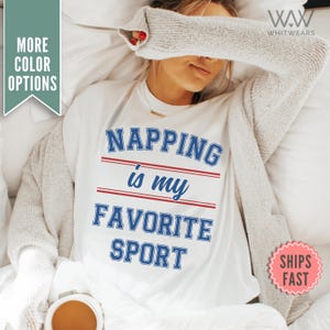 Könnte beinhalten: Weißes T-Shirt mit blauem und rotem Text, der "NAPPING is my FAVORITE SPORT" lautet.