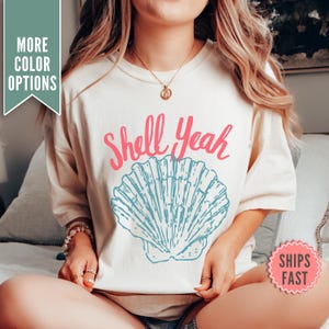 May include: Cream-colored t-shirt with the words "Shell Yeah" in pink script above a blue seashell graphic. The shirt has short sleeves and a relaxed fit. The image also includes a "More Color Options" tag and a "Ships Fast" sticker.