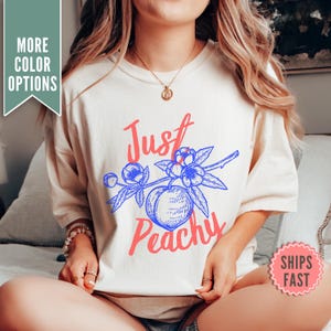 Puede incluir: Una camiseta blanca con un diseño gráfico azul y rosa. El gráfico presenta un melocotón con hojas y el texto "Just Peachy".
