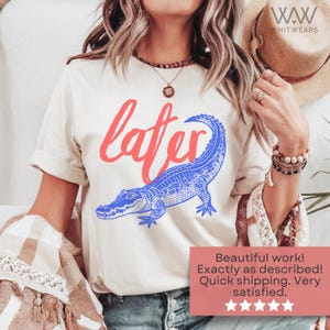 May include: A white t-shirt with a blue alligator and the word "later" in red. The alligator is facing left and has a detailed design.