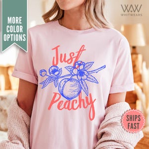 Peut inclure: Un t-shirt rose avec un motif graphique bleu et rouge qui dit "Just Peachy" avec une pêche et des fleurs.