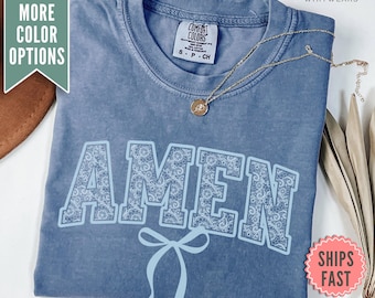 Può includere: Una maglietta blu con la parola "AMEN" in bianco con un motivo di pizzo blu chiaro. La parola è in un carattere grande e in grassetto. Un fiocco blu chiaro si trova sotto la parola.