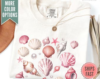 Op de afbeelding: Een beige T-shirt met een roze en wit schelpendesign. Het shirt heeft de tekst "Comfort Colors" op het label. Het shirt is gevouwen en heeft een gouden ketting met een hanger.