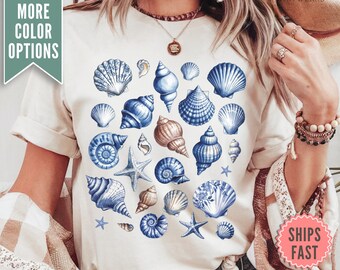 Op de afbeelding: Een wit T-shirt met een blauw en wit schelpmotief. Het shirt toont verschillende schelpen in verschillende maten en vormen, waaronder zeesterren.