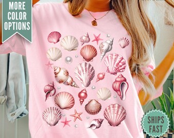 Op de afbeelding: Roze T-shirt met een patroon van schelpen, zeesterren en parels. De schelpen zijn in verschillende tinten roze en wit.