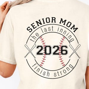 May include: Ivory-colored t-shirt with a baseball-themed graphic. The design includes the words "SENIOR MOM" and "2026" inside a baseball, with crossed bats. The text "the last inning" and "finish strong" are also included.