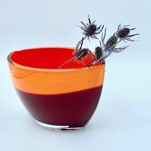 May include: A glass bowl with a layered color design. The top is bright red, transitioning to orange, and then to a deep burgundy at the base. The bowl holds several dried, spiky, purple-blue flowers. Home decor.