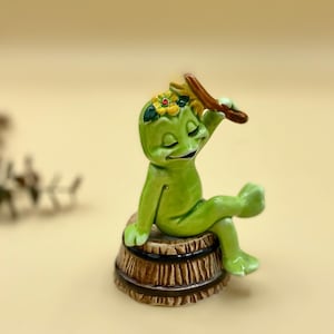 May include: A green frog figurine with a flower crown sits on a brown wooden barrel. The frog is holding a twig in its hand and has its eyes closed.