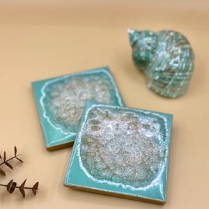 May include: Two square teal coasters with a white and gold agate design. The coasters have a light teal border.