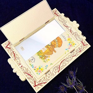 May include: A vintage ivory-colored compact mirror with a hinged lid. The frame features a leaf and branch design in red. The interior has a mirror and a colorful illustration with the word "Birthday!" and a floral design.