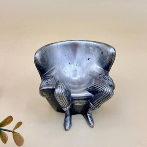 May include: A silver-toned figurine of a person sitting in an armchair. The figure has a metallic finish with dark shading, wearing striped trousers and shoes. The person is seated with hands resting on the armrests of the chair, creating a singular piece.