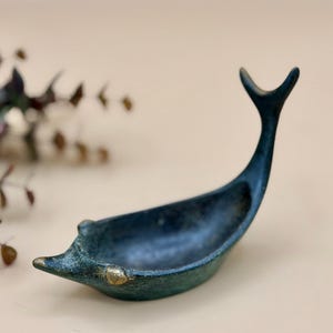 May include: A dark teal-colored, boat-shaped decorative dish with a curved tail and a small, gold-colored accent on the front. The dish has a textured surface and is likely made of metal. The design is reminiscent of a stylized whale or fish.