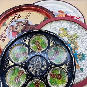 May include: A collection of vintage serving trays. The trays feature various designs, including illustrations of people, holiday scenes, and pine cones. The trays are in various colors, including red, black, and white. The trays are round and appear to be made of metal.