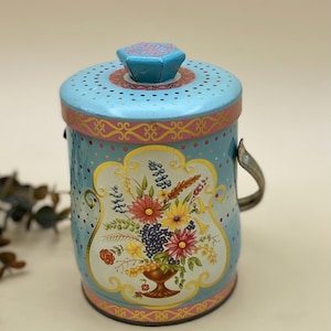 May include: A vintage-style, cylindrical metal container with a lid and handle. The container is light blue with pink and gold accents, and a floral design on the front. The lid has small holes and a decorative knob.