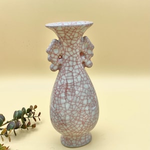 May include: A small, white ceramic vase with a crackle glaze and red accents. The vase has two handles on the sides and a round base.
