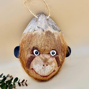 May include: A handcrafted monkey face ornament made from a coconut shell. The monkey has white eyes, black ears, and a smiling red mouth. The ornament is suspended by a natural fiber cord, perfect for hanging. The monkey's face is painted with brown and tan details.