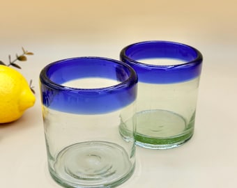 May include: Two clear drinking glasses with a vibrant blue rim. The glasses are cylindrical and appear to be made of glass. A yellow lemon is partially visible in the background, suggesting a refreshing beverage.