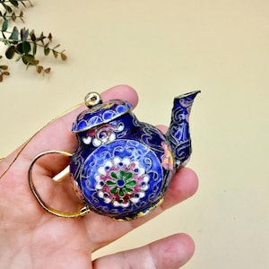 May include: A miniature blue teapot ornament with intricate floral designs in pink, green, and white. The teapot has a gold handle and spout, and a gold string for hanging. The ornament is held in a hand.