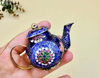 May include: A miniature blue teapot ornament with intricate floral designs in pink, green, and white. The teapot has a gold handle and spout, and a gold string for hanging. The ornament is held in a hand.