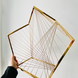 May include: A gold metal geometric sculpture with a square base and a series of intersecting lines forming a three-dimensional shape.