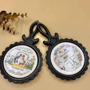 May include: A set of two black cast iron hot pads with white ceramic inserts. The inserts feature colorful embroidered designs with sayings. One pad says "Kissin' Don't Last Cookin' Do" and the other says "First the Toast Then the Roast Happy Hostess Happy Host".