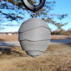 May include: A gray, 3D printed, egg-shaped ornament with a textured, layered surface. The ornament is hanging from a black hook.