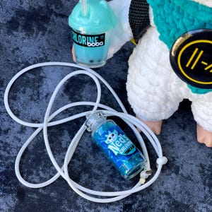 May include: A glass bottle pendant filled with blue glitter, inscribed with "Ned's Chlorine", on a white cord. A turquoise boba drink and a plush toy are also visible. The pendant is a unique accessory.