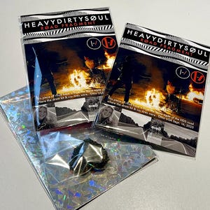 May include: Two packets labelled "HEAVYDIRTYSØUL ROAD FRAGMENT" with a black and white image of a fire. One packet contains a small, black, irregularly shaped object in a clear bag. The text on the packets reads "This piece of road base is from the loose edge of where the drum kit & car fires were - Ohio USA Sept. 16, 2025."