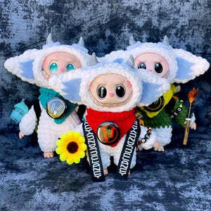 May include: Three plush dolls wearing white and blue hooded outfits, each with a different coloured scarf and accessory. One doll has a red scarf, another a teal scarf, and the third a yellow scarf. Each doll has a different accessory, including a sunflower and a small broom.