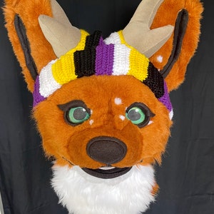 May include: A brown and white furry head with large ears and antlers. The head has green eyes and a black nose. A knitted headband in non-binary colours of yellow, black, purple, and white is on the head. The head has a white furry chin.