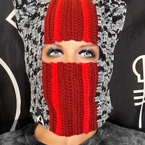 Könnte beinhalten: Eine handgefertigte Häkel-Balaclava in einem auffälligen Farbschema. Die Kopfbedeckung weist eine Mischung aus grauem und schwarzem gemustertem Häkelwerk mit einem kräftigen roten Mittelteil und roten Streifen auf. Die Sturmhaube hat Ohrenklappen und eine Gesichtsöffnung.