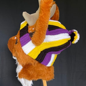 Op de afbeelding: Een bruine pluche elandkop met geweien, met een gehaakte muts en sjaal in de kleuren van de non-binaire pride-vlag: geel, wit, paars en zwart. De muts heeft een bijpassende pompon. De eland heeft witte vacht op zijn borst.