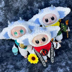 May include: Three plush dolls wearing white and blue horned hats and matching outfits. One doll has a red scarf and a black strap with white text. A yellow sunflower and a small blue cup are also present.