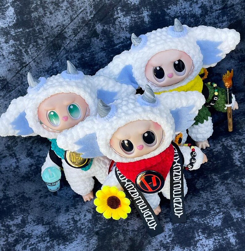May include: Three plush dolls wearing white and blue horned hats and matching outfits. One doll has a red scarf and a black strap with white text. A yellow sunflower and a small blue cup are also present.