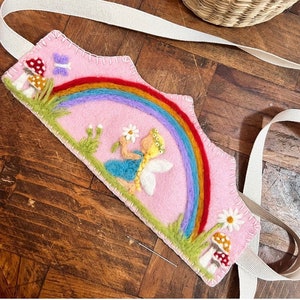 May include: A pink felt headband with a rainbow, a fairy, mushrooms, and flowers embroidered on it. The headband has white ribbon ties.