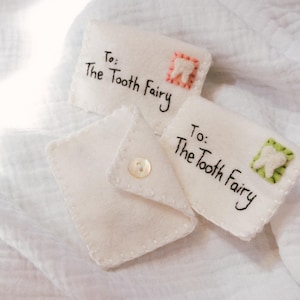 May include: Three small, white felt envelopes. Each has the text "To: The Tooth Fairy" in black script. Two envelopes feature a tooth graphic with red and green borders. One envelope is open, secured with a button.