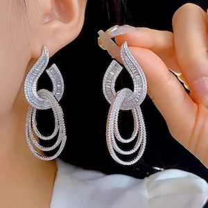 May include: A pair of silver-toned drop earrings featuring a flame-shaped top section encrusted with baguette and round clear stones. Below the top section, three interlocking oval-shaped hoops are suspended, each covered in small clear stones.